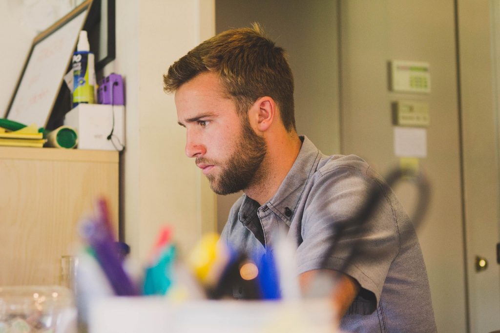 Ein junger Mann mit Bart konzentriert sich beim Arbeiten an seinem Schreibtisch, umgeben von Büromaterialien, symbolisierend einen Praktikanten während seines Studiums."